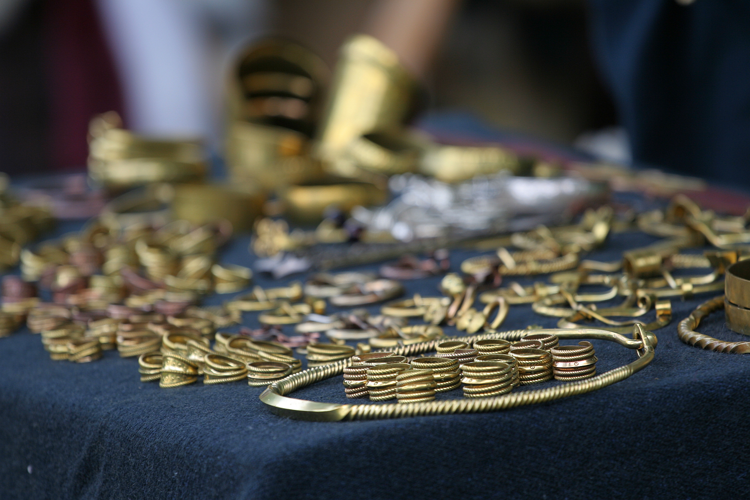 A close-up view of assorted gold jewelry pieces displayed on a dark cloth. The collection, reminiscent of the opulence seen at Kilkim Žaibu 2011, includes bracelets, rings, necklaces, and other ornamental items with intricate designs and metallic luster. Some pieces are arranged in rows; others are scattered, showcasing various sizes and shapes.