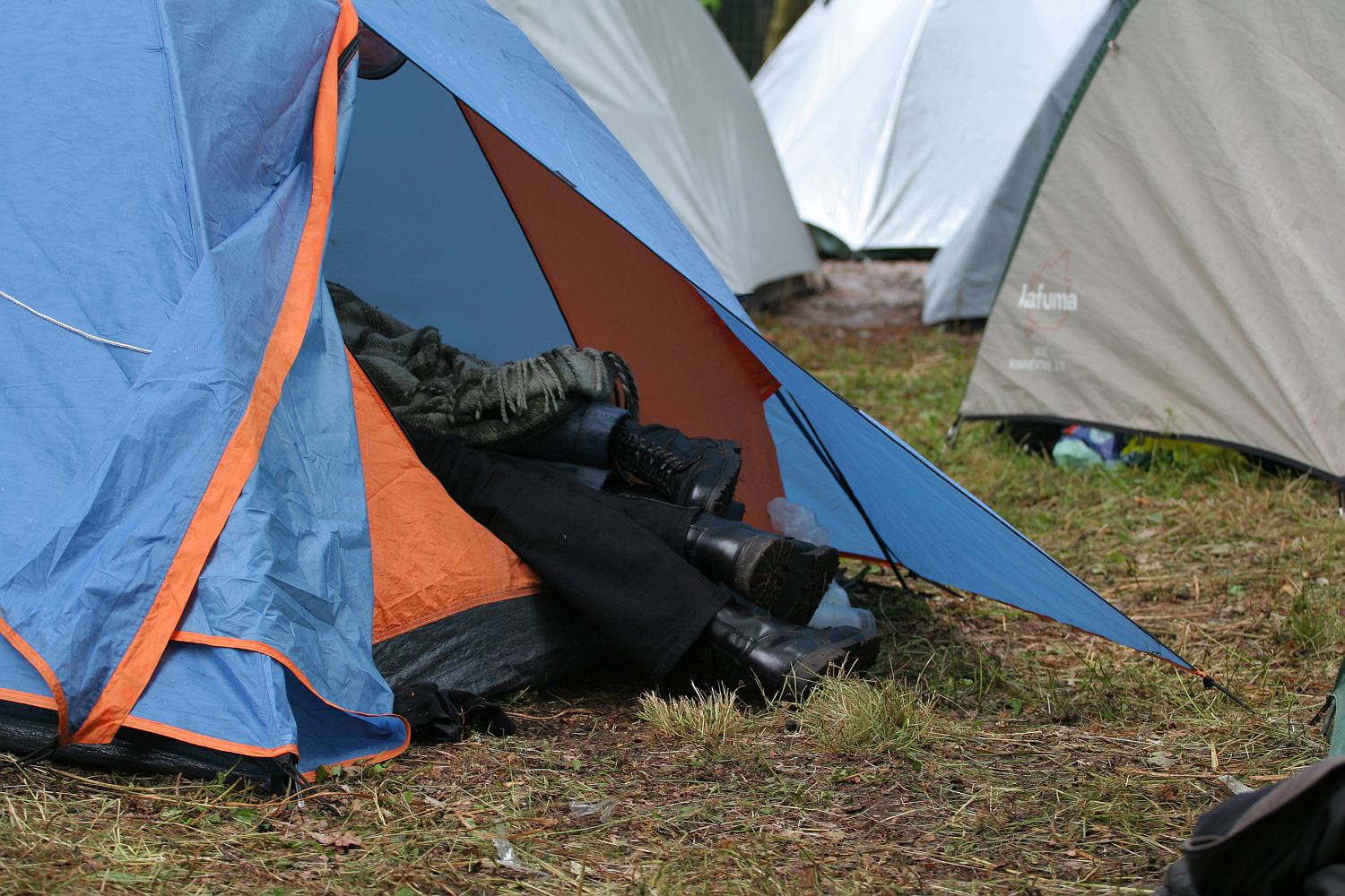 A blue and orange tent is set up on grassy ground. The tent flap is open, revealing three pairs of legs in black pants and black boots, partially covered by blankets. Other tents are set up in the background, suggesting a camping area at the 2011 Kilkim Žaibu festival with multiple tents.