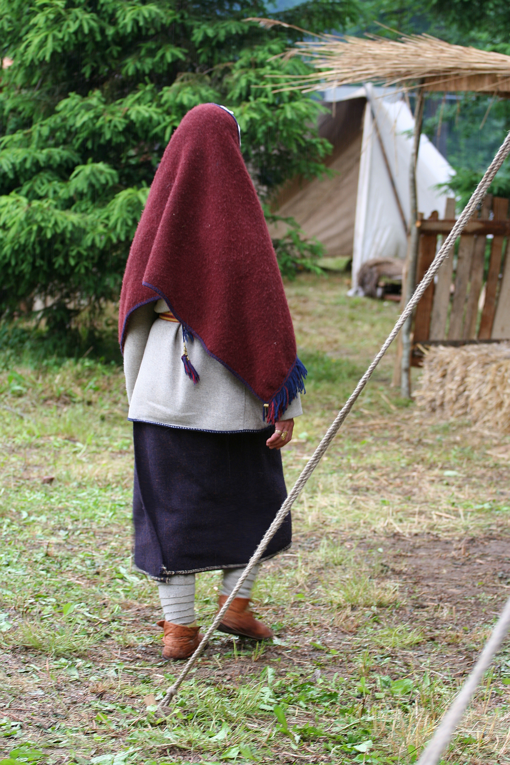 A person stands outdoors, covered head-to-toe in traditional clothing. They wear a deep red shawl over their head and shoulders, a white outer garment, a long dark skirt, and brown shoes. This scene from the 2011 Kilkim Žaibu festival features green trees, a rustic wooden fence, and a tent on grassy, slightly muddy ground.