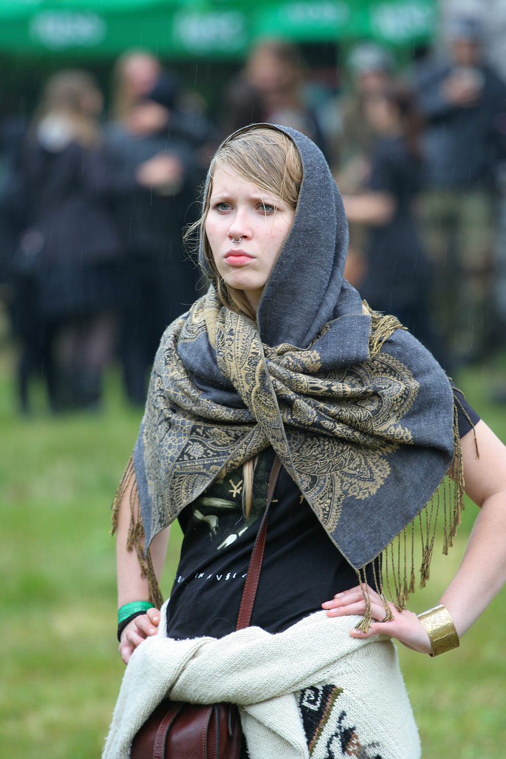 A young woman with blond hair stands outdoors in a grassy area during Kilkim Žaibu 2011. She wears a dark, intricately patterned shawl over her head and shoulders, a black shirt, and a white garment around her waist. With a serious expression, people can be seen in the blurred background under a green canopy.