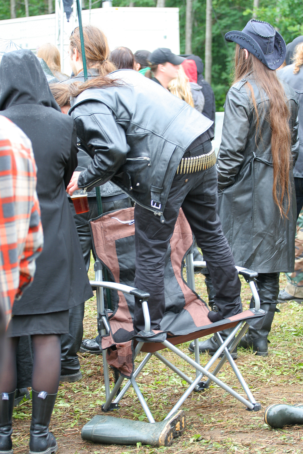 A person with long hair and a black leather jacket and pants stands on a folding camping chair at what appears to be the Kilkim Žaibu festival in 2011. The brown chair has armrests and a metal frame. Nearby, another person in a hooded jacket is visible, with more people in the background.