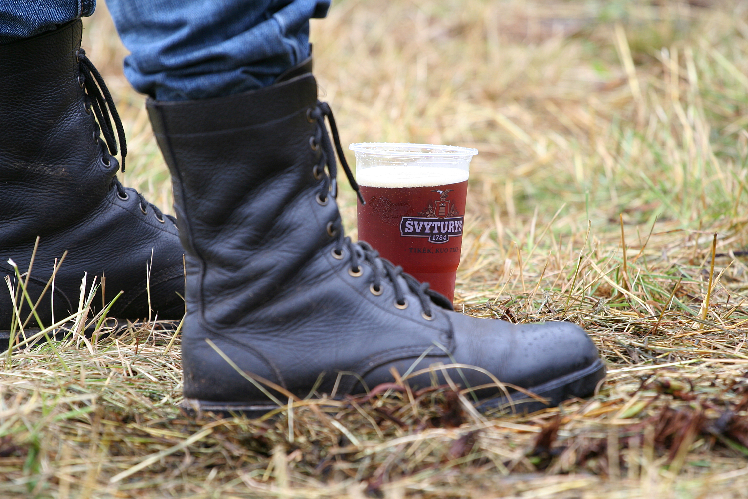 A close-up image of a person wearing black leather combat boots standing on grass at the Kilkim Žaibu festival. The individual is also clad in blue jeans. Beside the boots, there's a plastic cup filled with dark beer, crowned with white foam and bearing a visible label, capturing the essence of 2011 festivities.