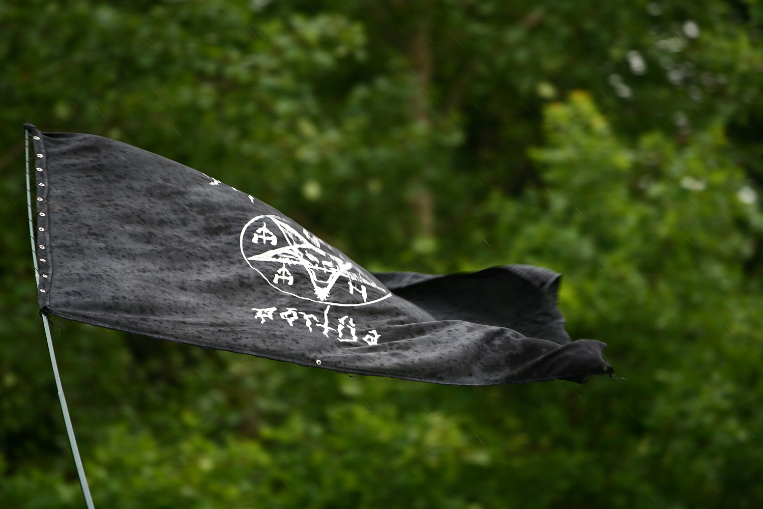 A black flag with white symbols and markings waves in the wind against a backdrop of lush green trees. The flag appears slightly wet, possibly from recent rain at Kilkim Žaibu in 2011, and is attached to a metal pole on the left side of the image. The symbols on the flag include geometric shapes and letters.