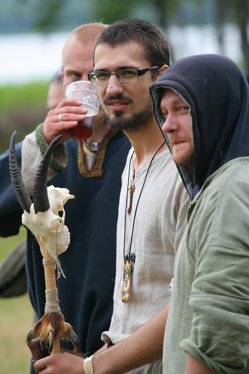 Three men are standing outdoors at Kilkim Žaibu 2011, dressed in medieval-inspired clothing. The man in the center has short dark hair, glasses, and a beard, and holds a staff adorned with a goat skull and horns. The man on the left is drinking from a cup, while the man on the right wears a hooded garment and has a light beard.