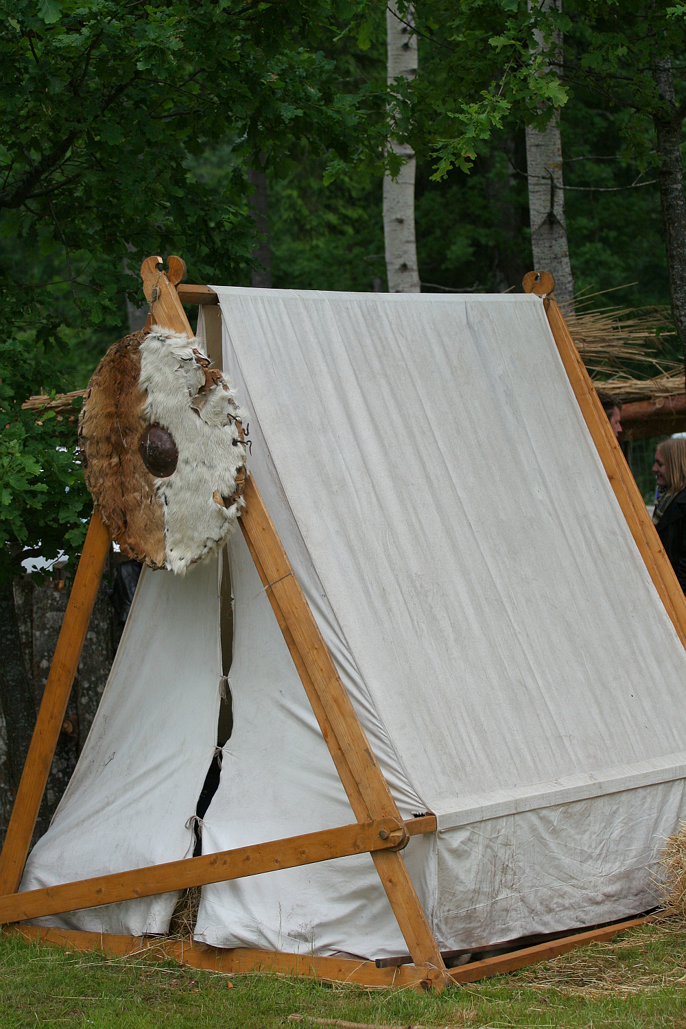 A small triangular canvas tent with wooden supports is set up in a forested area, reminiscent of the Kilkim Žaibu 2011 festival. A round shield with fur and leather adorns the left side of the tent. The background includes trees and partial glimpses of other tents or structures among the greenery.