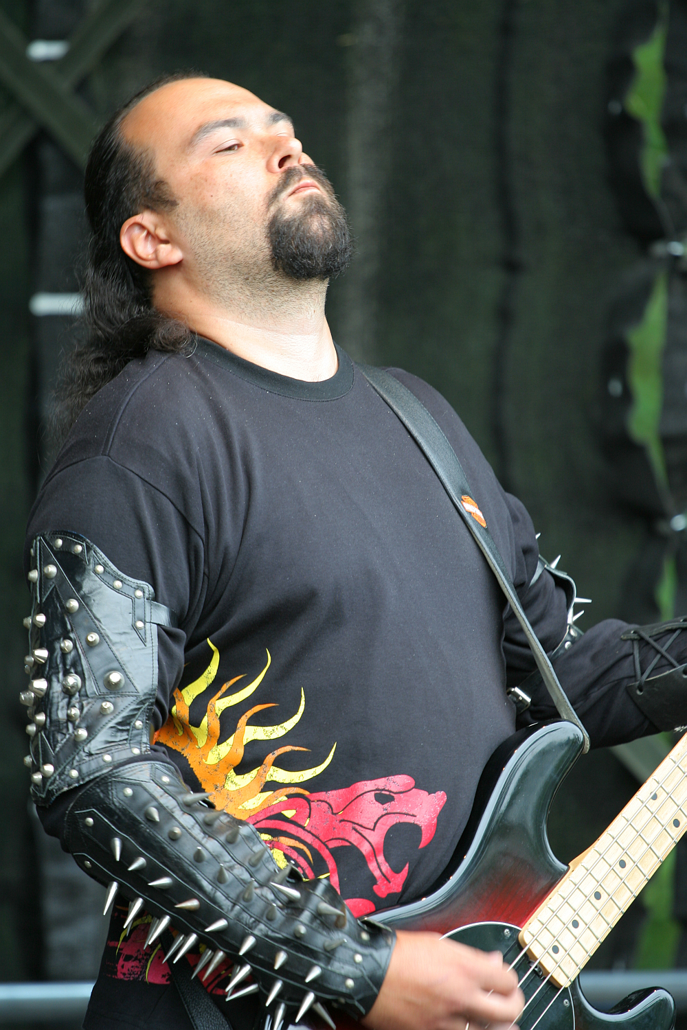 A person with long dark hair, a beard, and mustache is playing a black electric guitar. They are wearing a black shirt with a red and yellow sun design and black armbands adorned with metal spikes. Their eyes are closed as they appear deeply absorbed in the music, reminiscent of Kilkim Žaibu 2011.