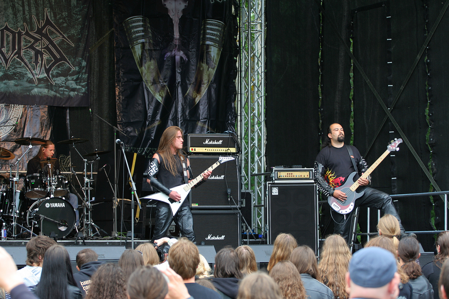Three musicians perform on an outdoor stage at the 2011 Kilkim Žaibu festival. The guitarist on the left plays a Flying V guitar while the bassist on the right plays a bass guitar. A drummer is visible behind them. The audience, with long hair, stands close to the stage amidst amplifiers and a backdrop with the band logo.
