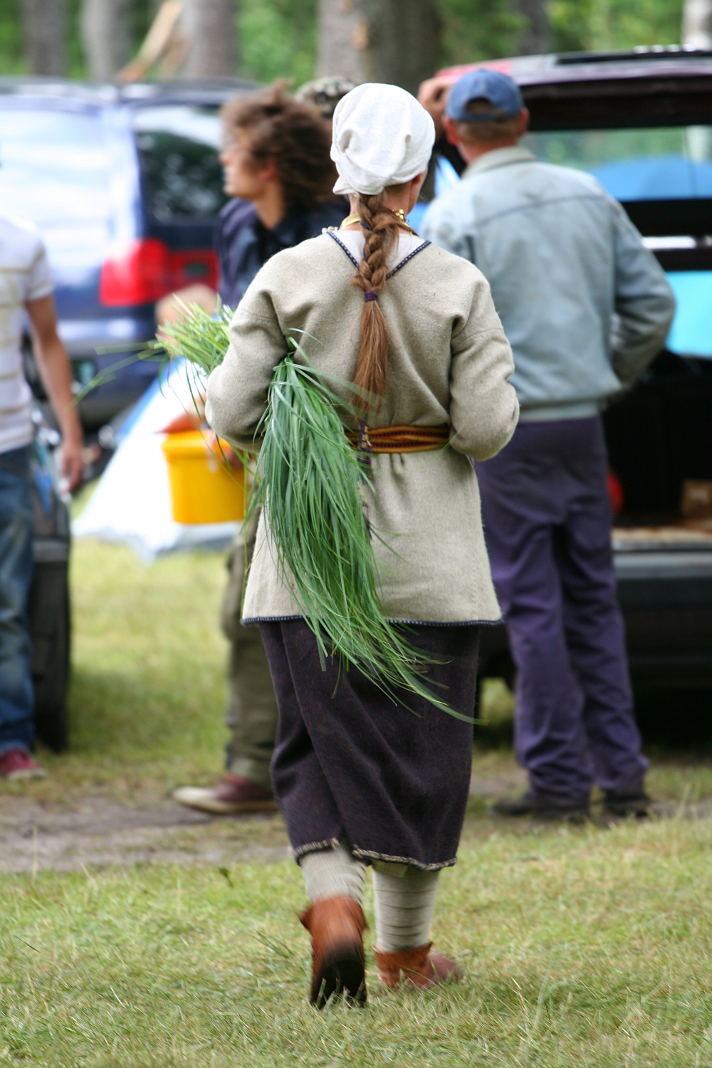 A person with long braided hair, wearing a beige shawl, grey dress, and dark apron, is seen from behind holding a large bunch of green herbs. They walk on grass towards a car with its trunk open, surrounded by others who are in casual attire and appear to be engaging in an outdoor activity reminiscent of Kilkim Žaibu 2011.