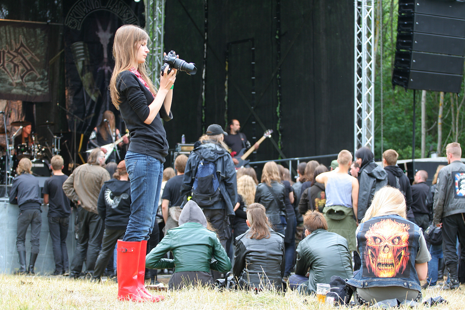 At the 2011 Kilkim Žaibu festival, a young woman with long brown hair, dressed in a black outfit and red rain boots, holds a camera and faces the stage. The crowd, some seated and others standing, watches the band perform amid trees and stage equipment. One person wears a vest with a skeletal face design.