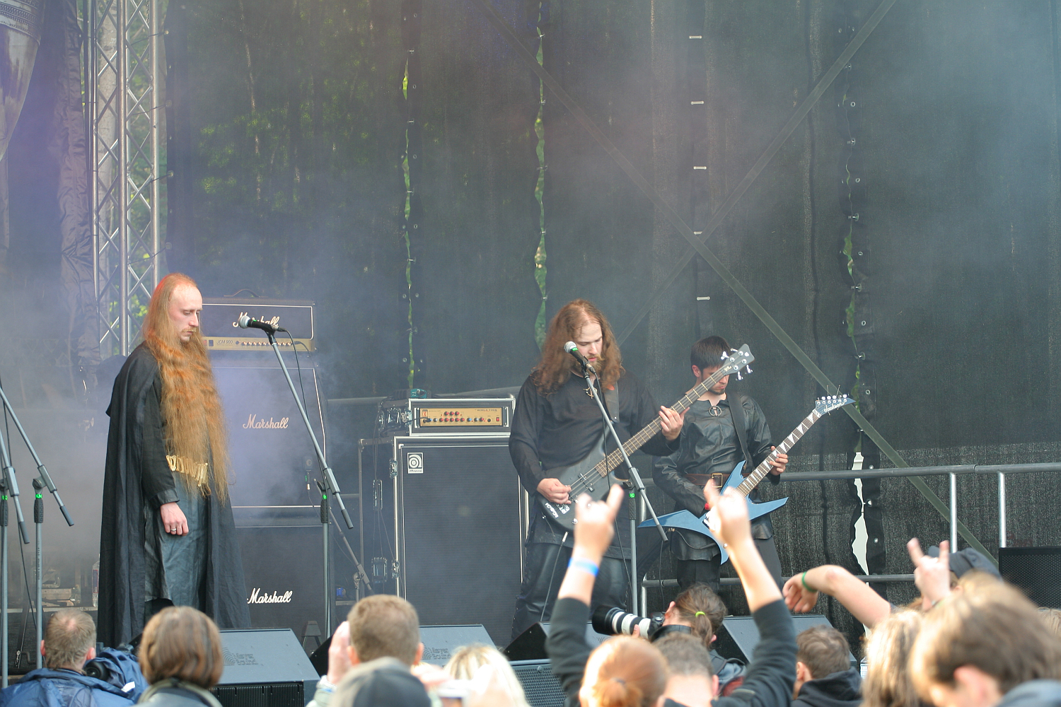 At the 2011 Kilkim Žaibu festival, a heavy metal band performs on an outdoor stage with fog effects. Three members play electric guitars and bass, wearing dark clothing, while an audience watches and raises their hands in appreciation. Amplifiers with the "Marshall" logo can be seen in the background.
