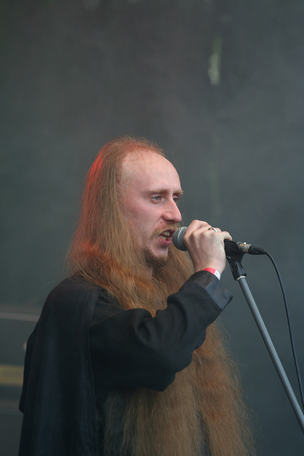 A person with long, flowing hair and a receding hairline is singing into a microphone on a stand. They are wearing a dark shirt and standing against a smoky, darkened background, likely on a stage at the 2011 Kilkim Žaibu festival. The lighting casts a slight reddish hue on their hair and face.