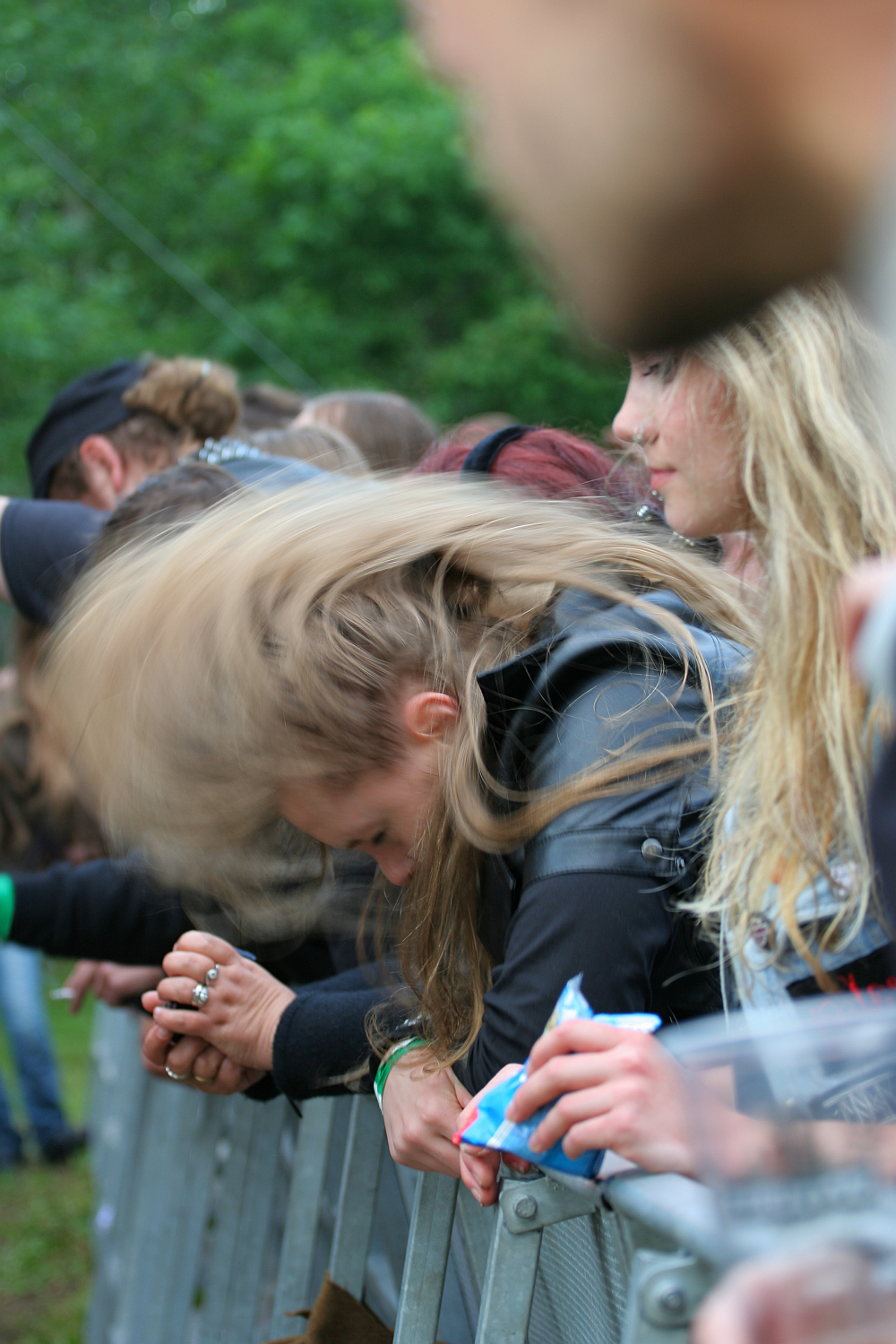 Several people stand in a row behind a metal barricade at an outdoor event. The person in the foreground has long hair flipping forward, likely headbanging. They are holding onto the barricade. Trees and greenery can be seen in the background, suggesting Kilkim Žaibu 2011 is taking place in a park.