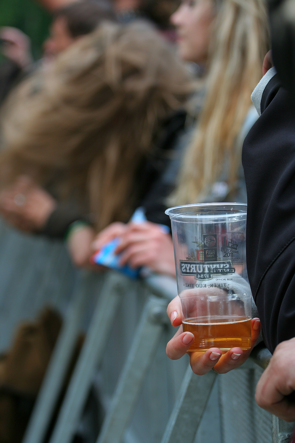 A close-up shot of a crowd at the 2011 Kilkim Žaibu festival. The focus is on a person holding a plastic cup with some beverage, possibly beer, inside it. Nearby, another person has long, blond hair flowing in the wind. Both are leaning against a metal barricade.