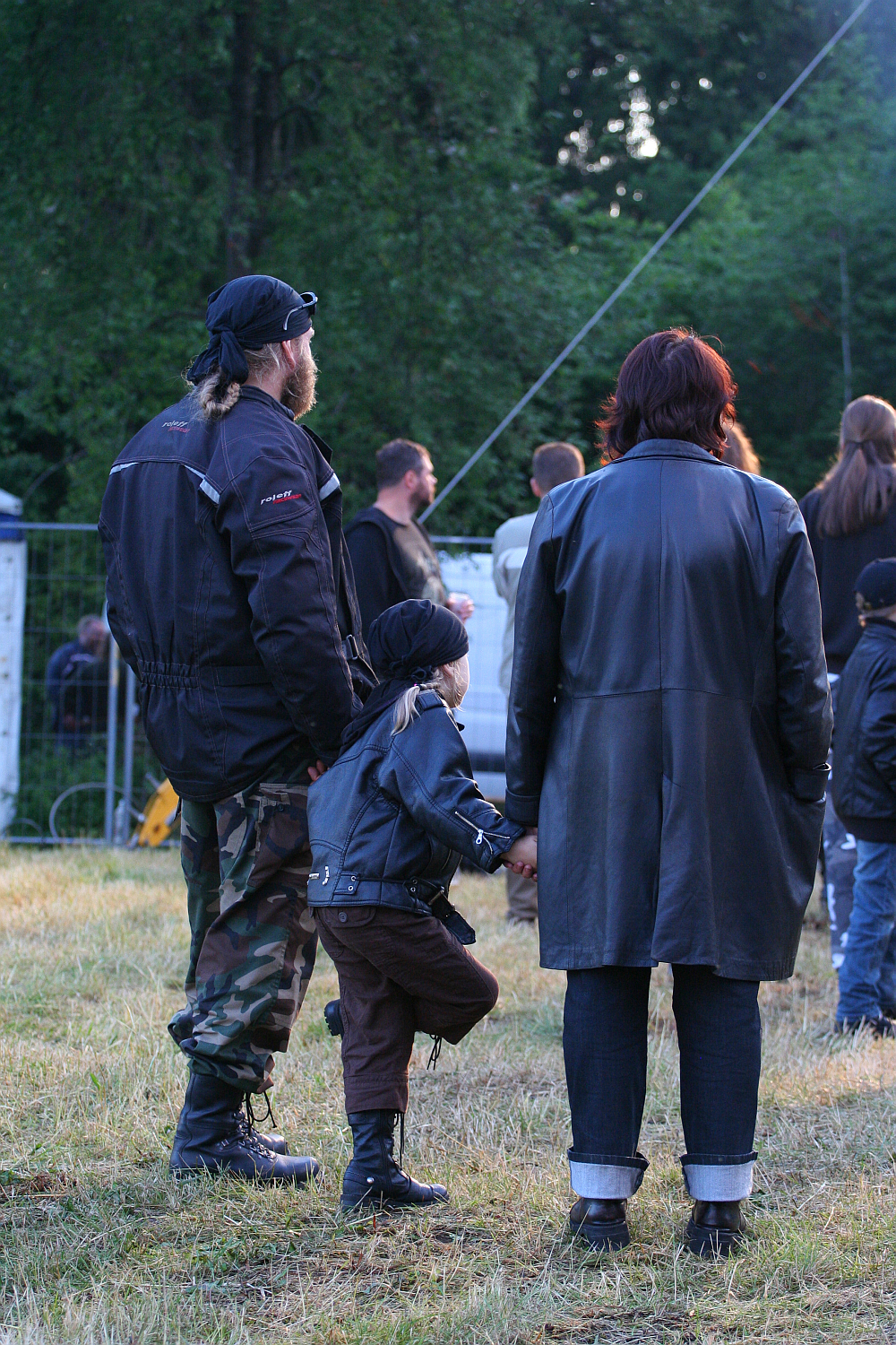 At the 2011 Kilkim Žaibu festival, a man, woman, and child, all in black jackets, stand together at an outdoor event. The man sports a bandana and camouflage pants; the woman has short hair and rolled-up jeans. The child, also wearing a bandana, holds the woman's hand while stepping on one foot. Trees and other attendees create a lively backdrop.