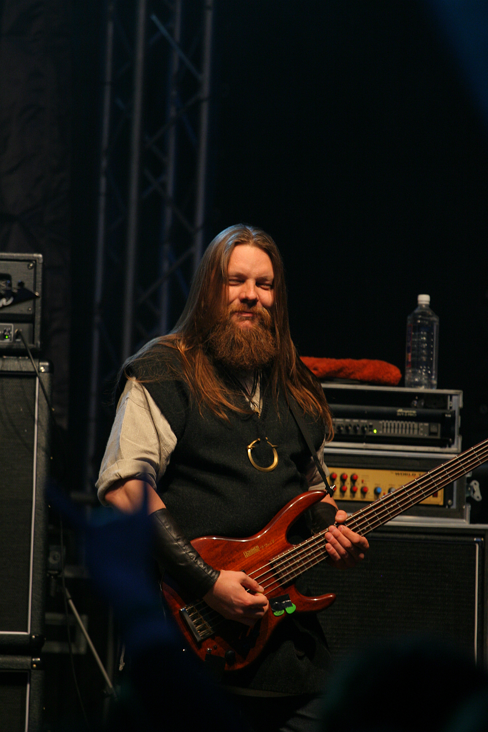 A man with long hair and a beard stands on stage, smiling while holding a brown electric bass guitar at Kilkim Žaibu 2011. He is dressed in dark clothing and wears a large ring necklace. Behind him, audio equipment and a water bottle rest atop an amplifier, with stage lighting and a partially visible crowd in the foreground.