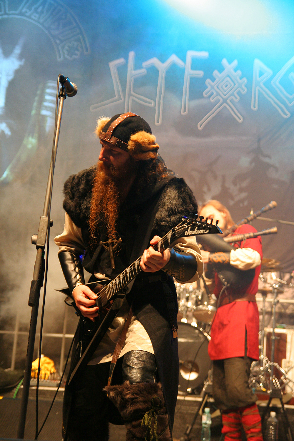 A musician with a long red beard, dressed in a Viking-inspired outfit, plays an electric guitar on stage. Behind him, another performer in a red and black costume plays drums. The stage backdrop features the name "Skálmöld" with Norse motifs. Smoke and blue lighting enhance the dramatic setting at Kilkim Žaibu 2011.