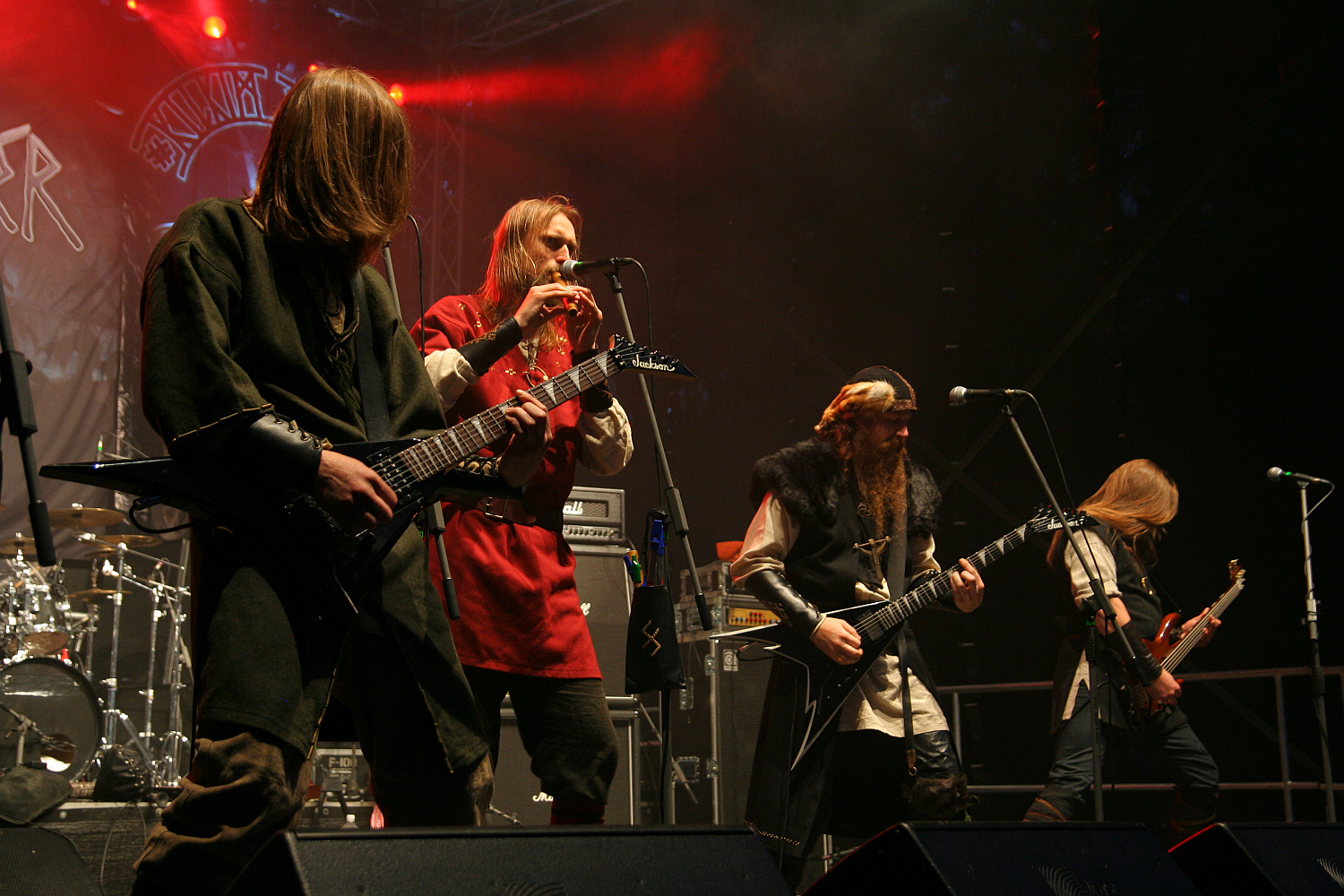 Musicians perform on a dimly lit stage at the 2011 Kilkim Žaibu festival. Four guitarists are playing: one in black, one in green, one in red (also playing a flute), and one in black with a fur collar. Amplifiers and drums stand visible in the background, accompanied by a backdrop of intricate designs.