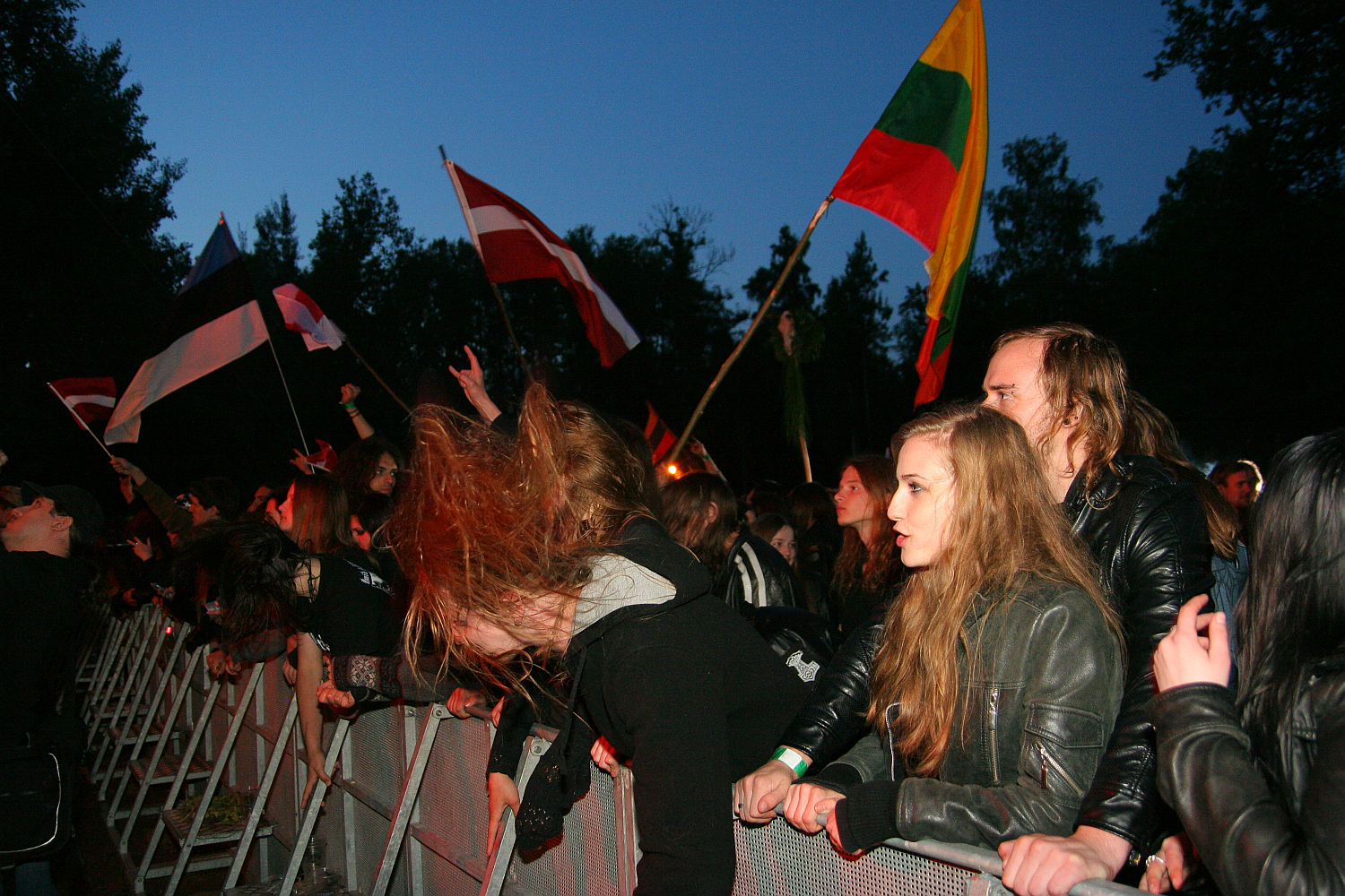 A crowd at the 2011 Kilkim Žaibu festival enthusiastically listens to live music. Attendees, many with long hair and dressed in dark clothing, lean against a metal barricade, some headbanging. Several flags, including the Lithuanian flag, are held aloft amidst the dusk setting with trees in the background.