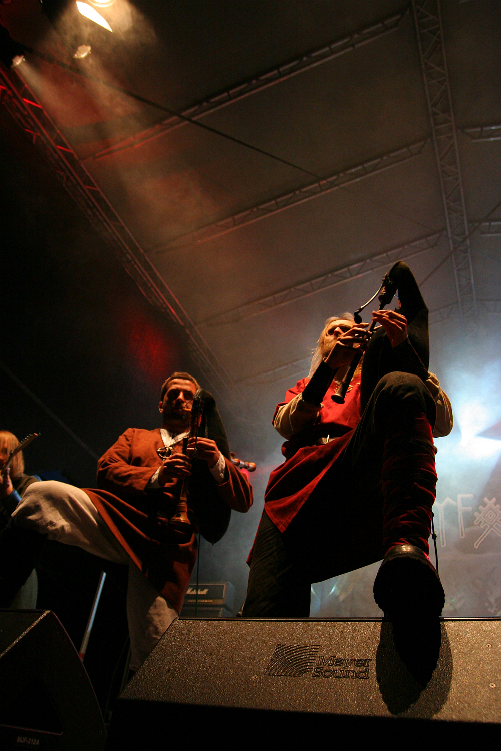 At the 2011 Kilkim Žaibu festival, two musicians perform on stage under dim lighting. The central focus is on one musician playing bagpipes and wearing a red tunic, while the other, also playing bagpipes, is dressed in a brown outfit. Smoke and colorful stage lights create a dramatic atmosphere. The audience is not visible.