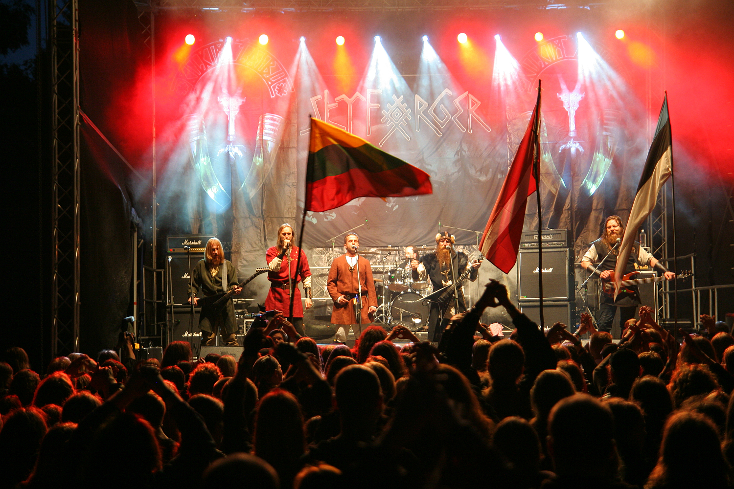 A rock band performs on stage with a large, enthusiastic crowd in the foreground at Kilkim Žaibu 2011 festival. The stage is illuminated with colored lights, and flags from various countries are being waved by audience members. The band's backdrop features their logo in bold letters.