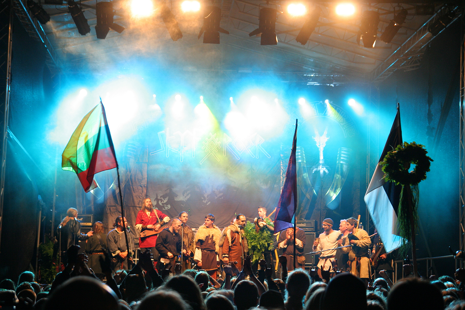 A vibrant concert stage at the 2011 Kilkim Žaibu festival features performers in medieval costumes holding various national flags. Bright lights and fog create a dramatic atmosphere. The audience, visible in the foreground, watches as the performers stand in a row, some raising their hands or flags in celebration.