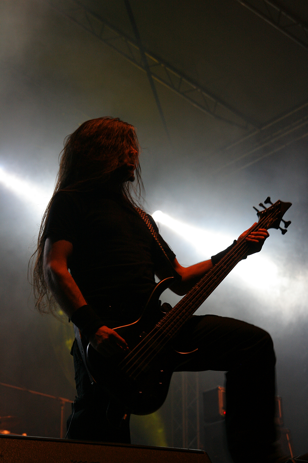 A musician with long, flowing hair plays a black electric guitar on stage at the 2011 Kilkim Žaibu festival. He is dressed in all black and surrounded by dramatic stage lighting, creating a high-contrast silhouette effect. Smoke and bright beams of light in the background add to the atmospheric, concert-like setting.