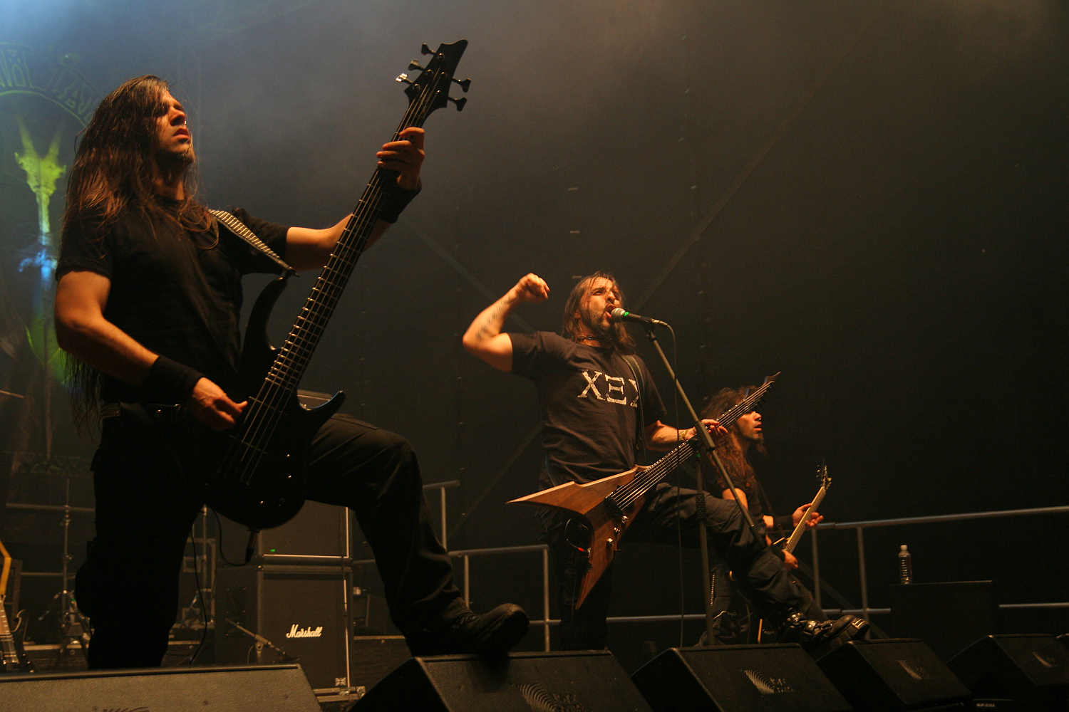At the 2011 Kilkim Žaibu festival, a heavy metal band performs on stage, featuring three members. The bassist on the left plays with intensity, the lead vocalist in the center shouts into a microphone while raising his fist, and the guitarist on the right focuses on his instrument. The stage is dimly lit, with amps and equipment visible in the background.