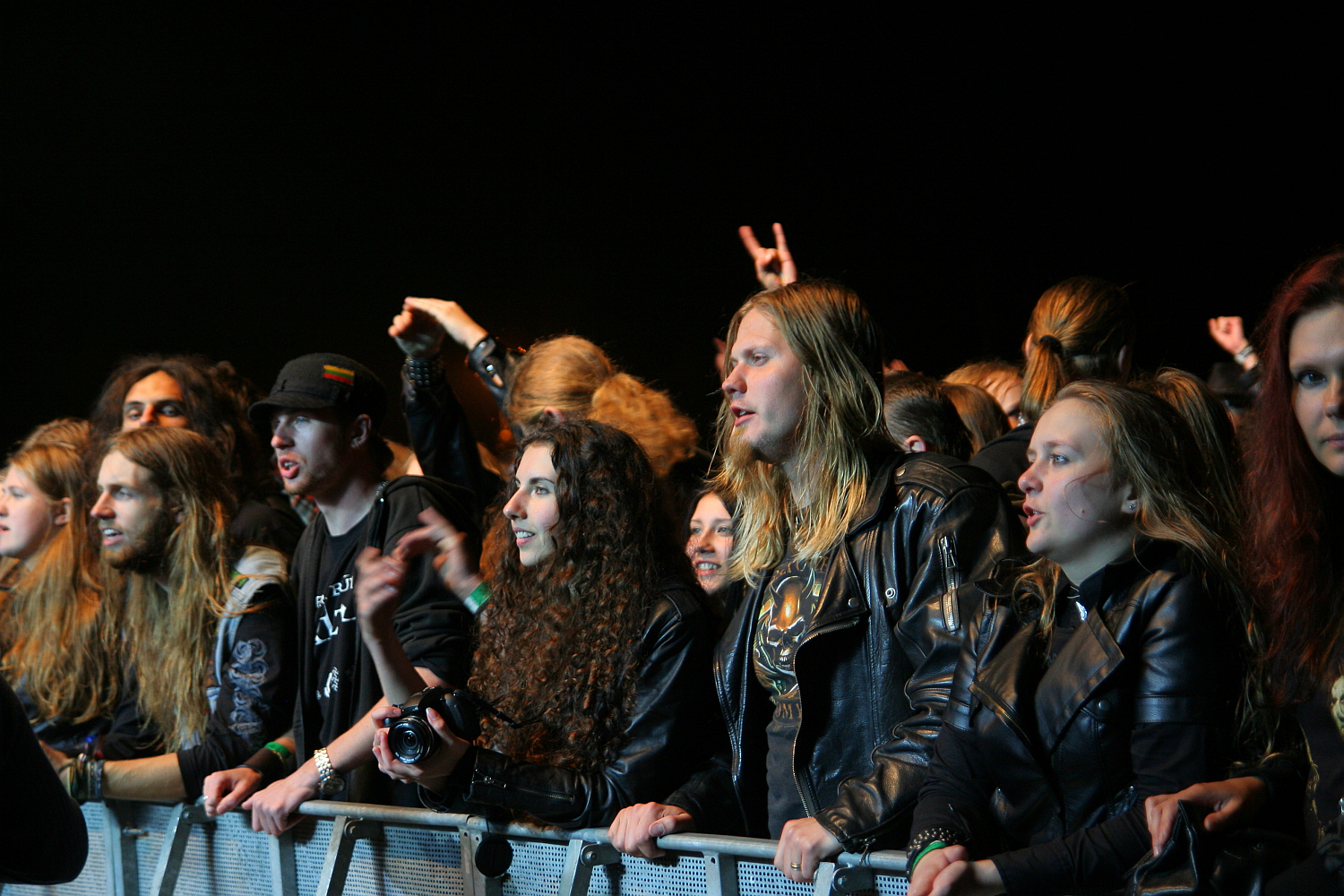 Concert-goers with long hair and leather jackets stand behind a metal barricade, enthusiastically watching a performance at the Kilkim Žaibu festival in 2011. Some have their hands raised, and one person is holding a camera. The dark background emphasizes the crowd's excited expressions and gestures.