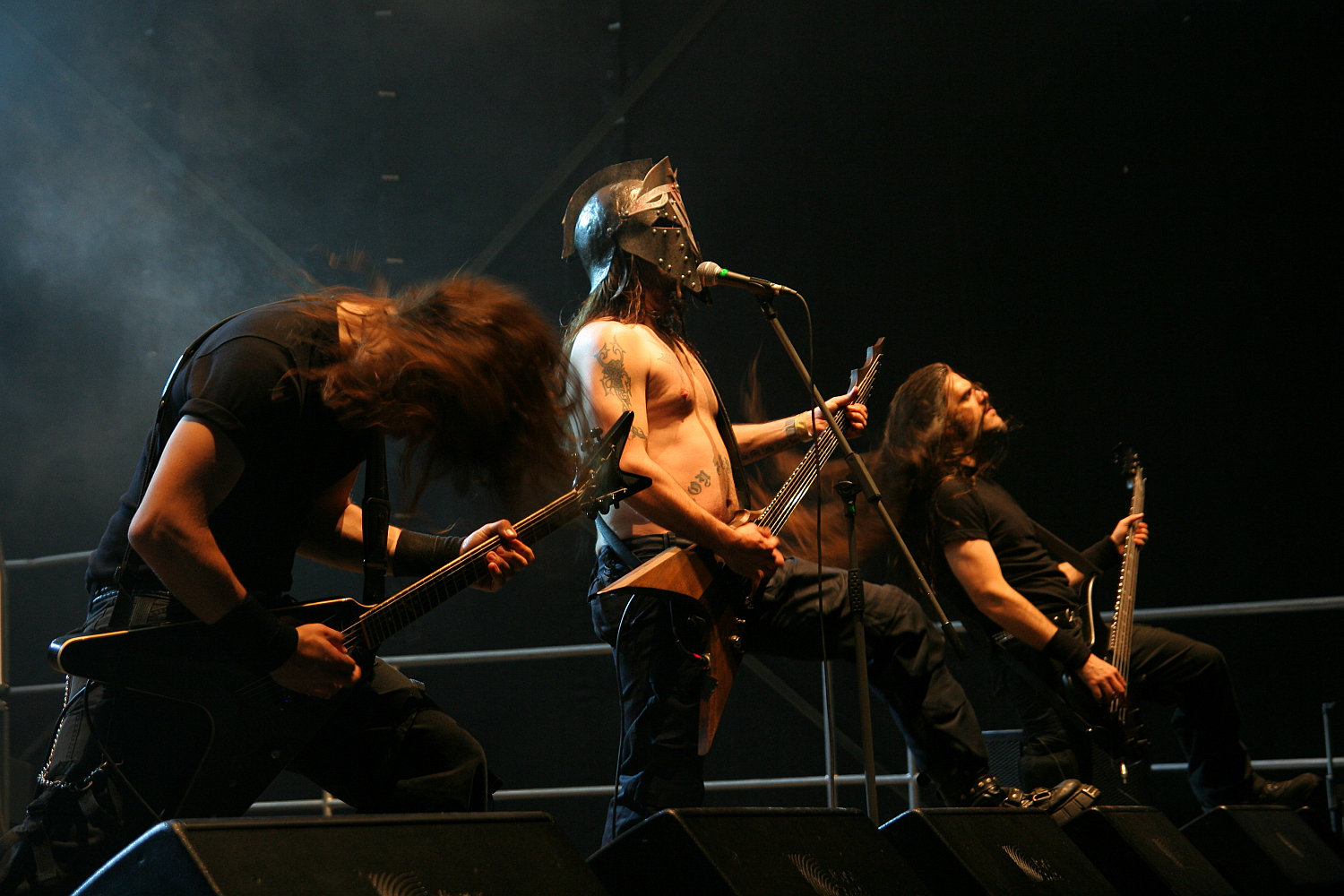 At Kilkim Žaibu 2011, four musicians perform energetically on stage. The shirtless lead singer wears a helmet and sings into a microphone. All band members have long hair and play electric guitars. The stage background is dark with stage lights illuminating the performers from above, creating a dramatic effect.