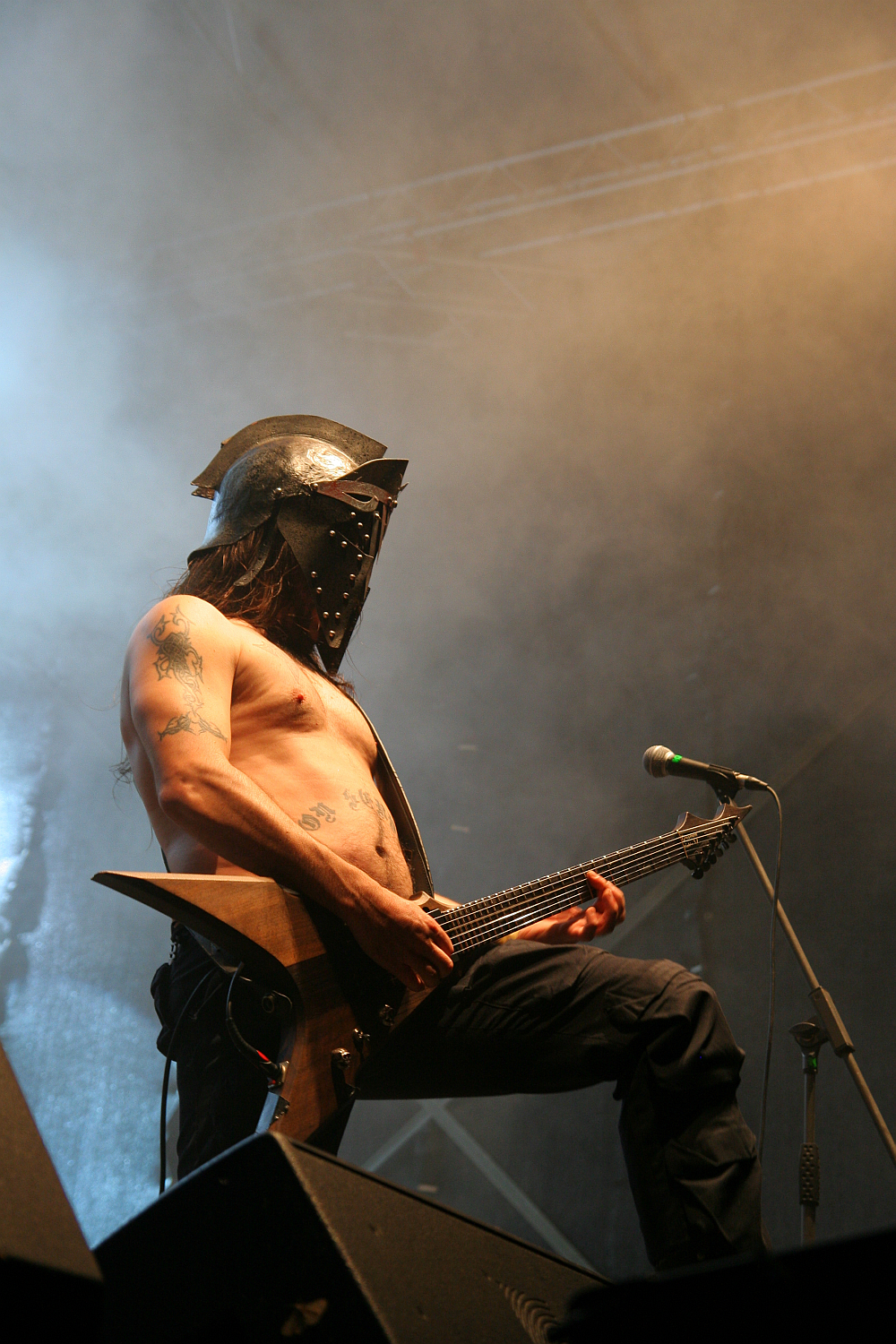 A shirtless musician wearing a metallic helmet with horn-like protrusions plays an electric guitar on stage at the 2011 Kilkim Žaibu festival. The stage is filled with a smoky haze, and microphones are positioned in front of him, while bright stage lights and equipment form the backdrop.