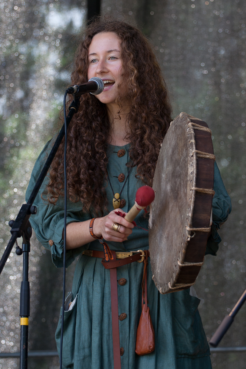 A woman with long curly hair, wearing a green dress with wooden buttons, performs on stage at Kilkim Žaibu 2012. She is holding a large, round hand drum and a red-tipped drumstick. Standing in front of a microphone, she smiles as she sings or speaks. The background is speckled with light.
