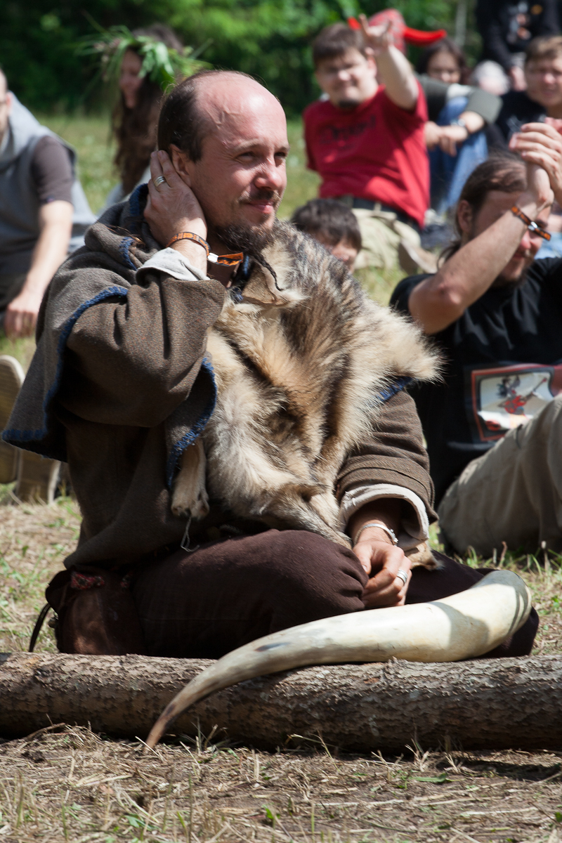 A man sits on the ground wearing medieval-style clothing with a fur draped over his shoulders. He holds one hand to his face and has a thoughtful expression. Surrounded by others, some clapping, the scene appears to be from the 2012 Kilkim Žaibu festival, set in a grassy reenactment area outdoors.