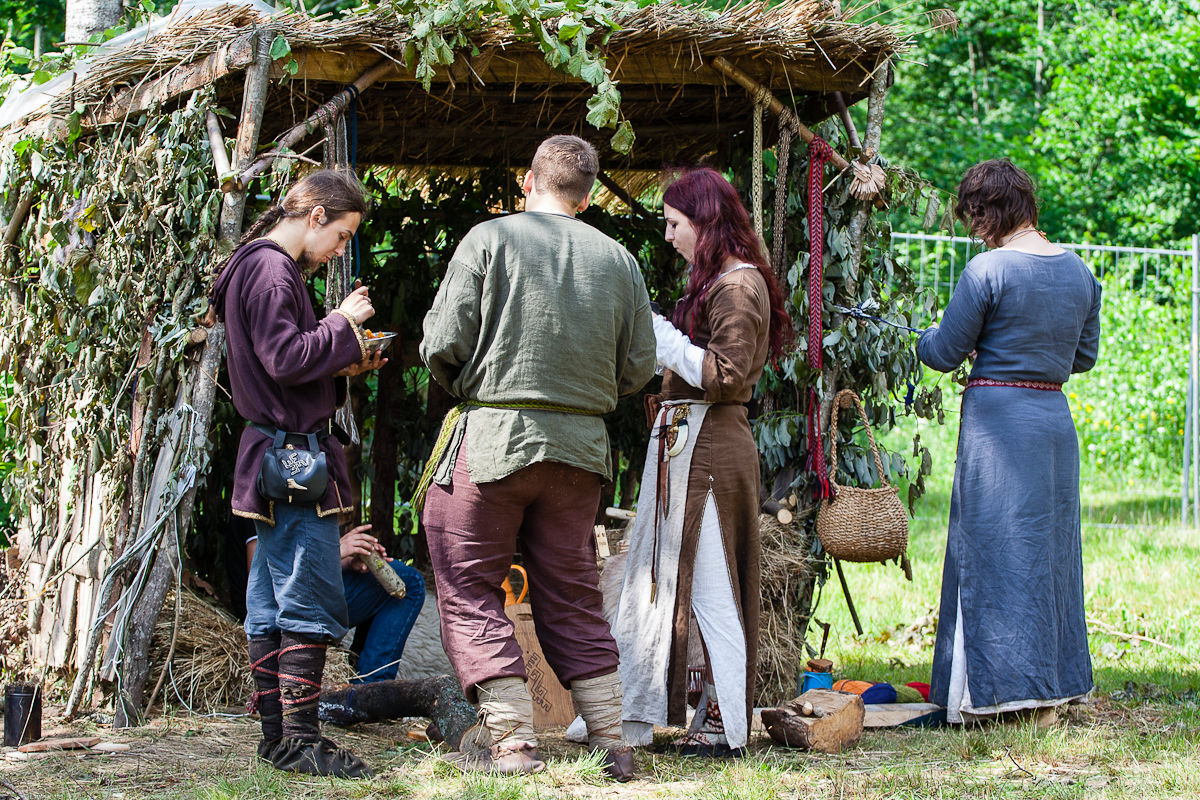 A group of five people in medieval attire stands and sits by a rustic shelter made of branches and straw in a lush, green outdoor setting at the 2012 Kilkim Žaibu festival. They appear to be engaged in various activities, with some looking down at objects in their hands or sitting inside the shelter.