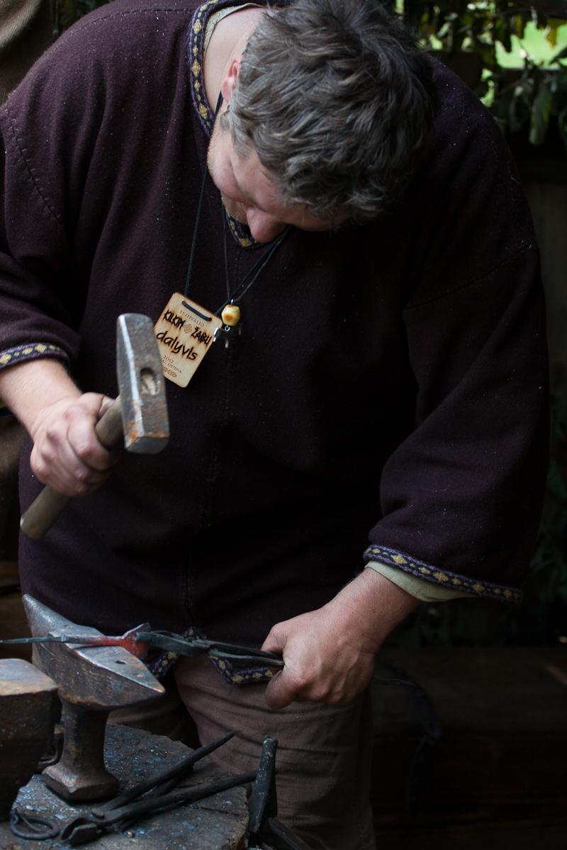 A person with short hair wearing a dark brown long-sleeved shirt is hammering red-hot metal on an anvil. They have a name tag hanging around their neck, likely from the Kilkim Žaibu festival. The background is slightly out of focus, suggesting an outdoor setting with greenery and tools alongside metalworking equipment are visible nearby.