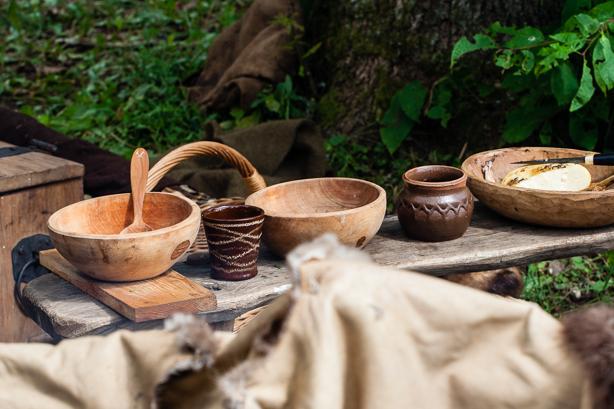 A rustic outdoor scene from the 2012 Kilkim Žaibu festival features a wooden table covered with various wooden and clay bowls, cups, and utensils. The background shows greenery and burlap sacks, suggesting a natural and simple setting. The artisanal bowls and cups are filled with food items.