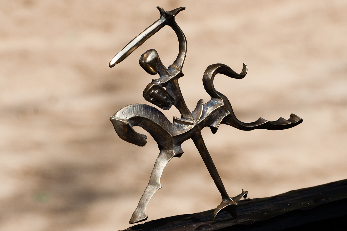A bronze sculpture depicts an abstract knight on horseback, raising a sword overhead. The minimalist design captures motion and energy, with fluid, exaggerated forms. Set against a softly blurred background, the dynamic and expressive figure channels the spirit of the Kilkim Žaibu festival in 2012.