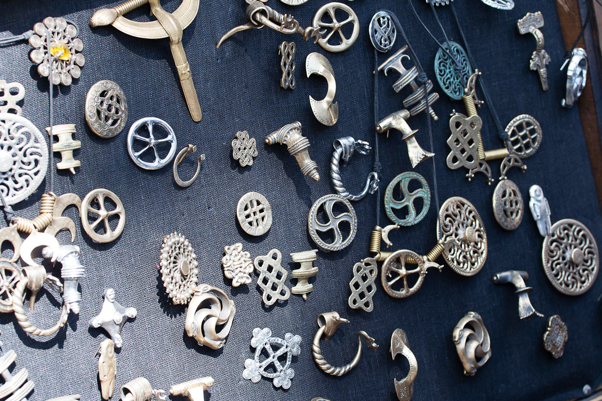 A display of numerous metal jewelry pieces is spread on a dark fabric surface at the 2012 Kilkim Žaibu festival. Various designs include intricate patterns, some resembling knots, spirals, and geometric shapes. The items vary in color, with some appearing silver and others gold. The arrangement is random yet visually appealing.