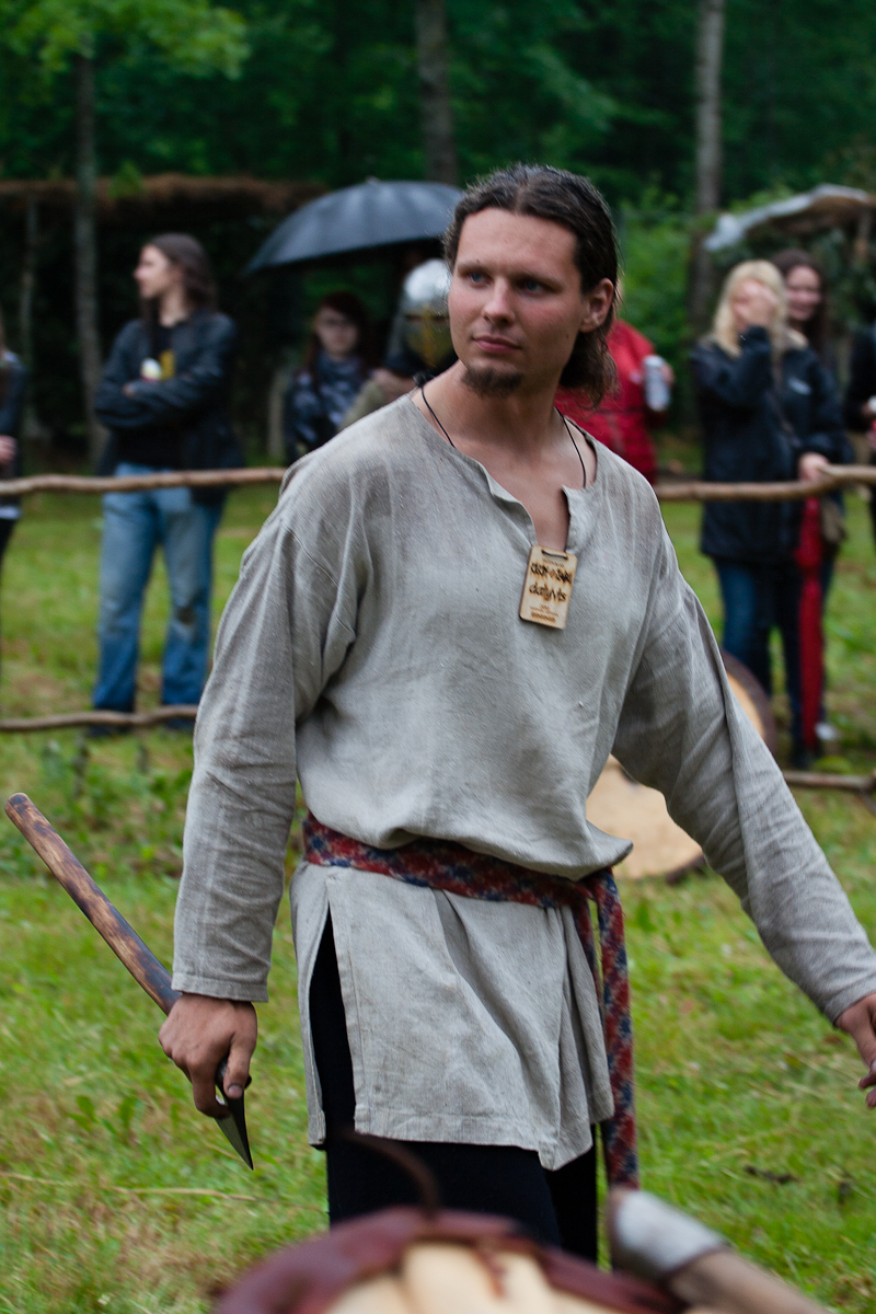 A man dressed in medieval attire, including a loose, beige tunic and a red patterned belt, stands on grass at Kilkim Žaibu 2012. He holds a wooden tool and wears a rectangular wooden pendant. Blurred spectators with modern clothing and umbrellas stand behind a wooden fence, with a forested background.