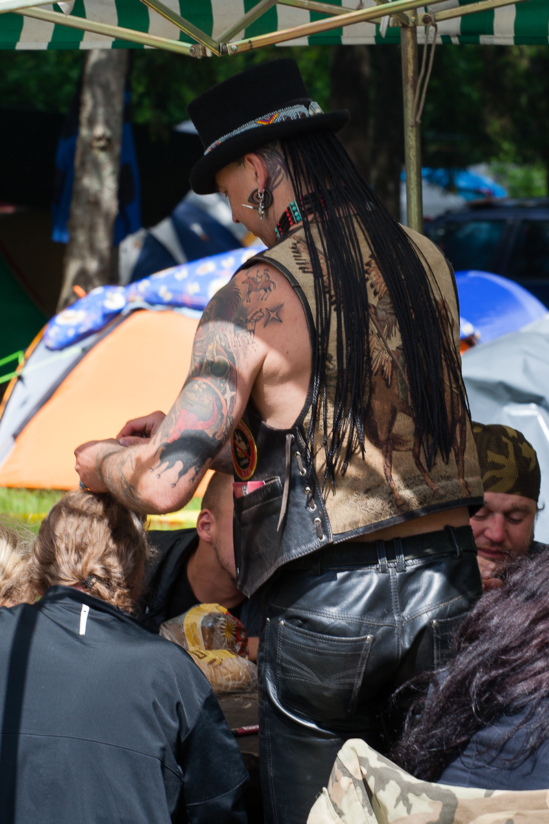A person with long dreadlocks, a top hat, and detailed tattoos on their arms leans over a group seated at a table outdoors. Sporting a sleeveless vest with a Native American motif and black leather pants, they evoke the wild spirit of Kilkim Žaibu 2012. Tents and trees in the background suggest a campsite setting.