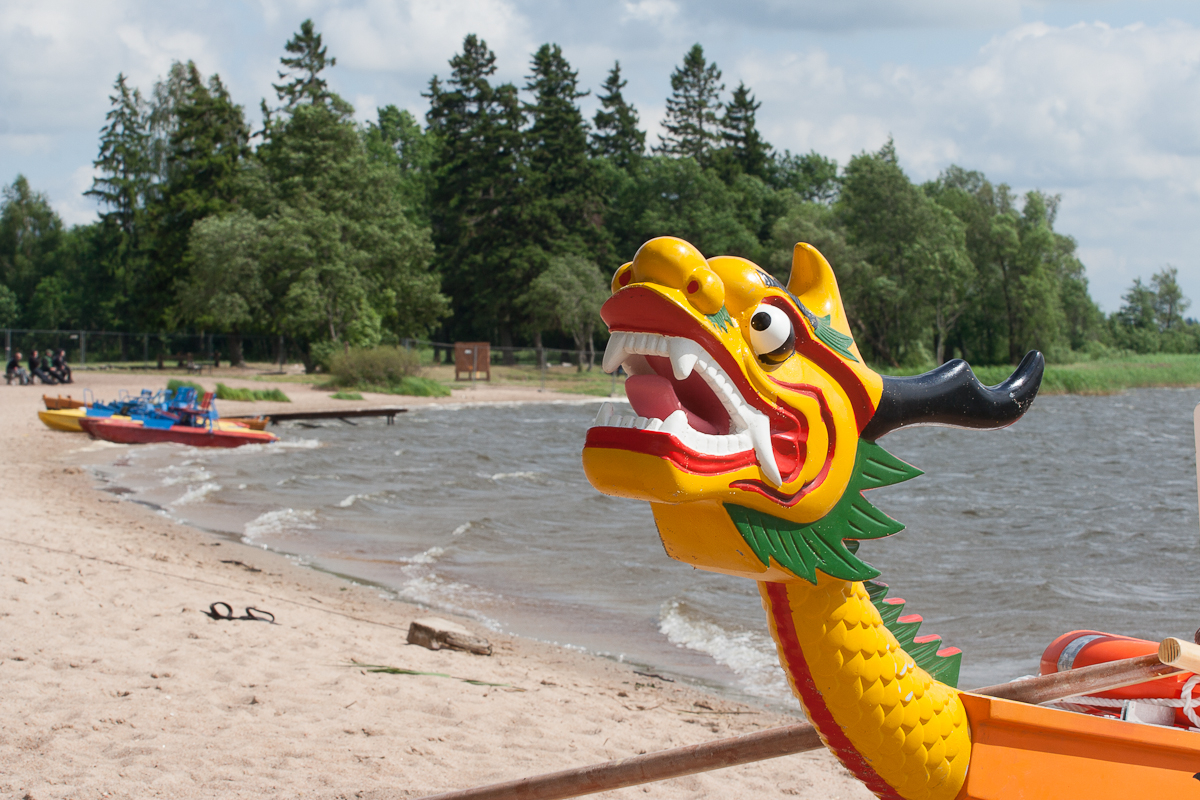 A brightly painted dragon boat with a yellow, red, and green dragon head rests on a sandy beach by a lake during the lively 2012 Kilkim Žaibu festival. Several other colorful boats are in the background near the shoreline. Tall trees and a partly cloudy sky are visible in the distance, completing this vivid scene.