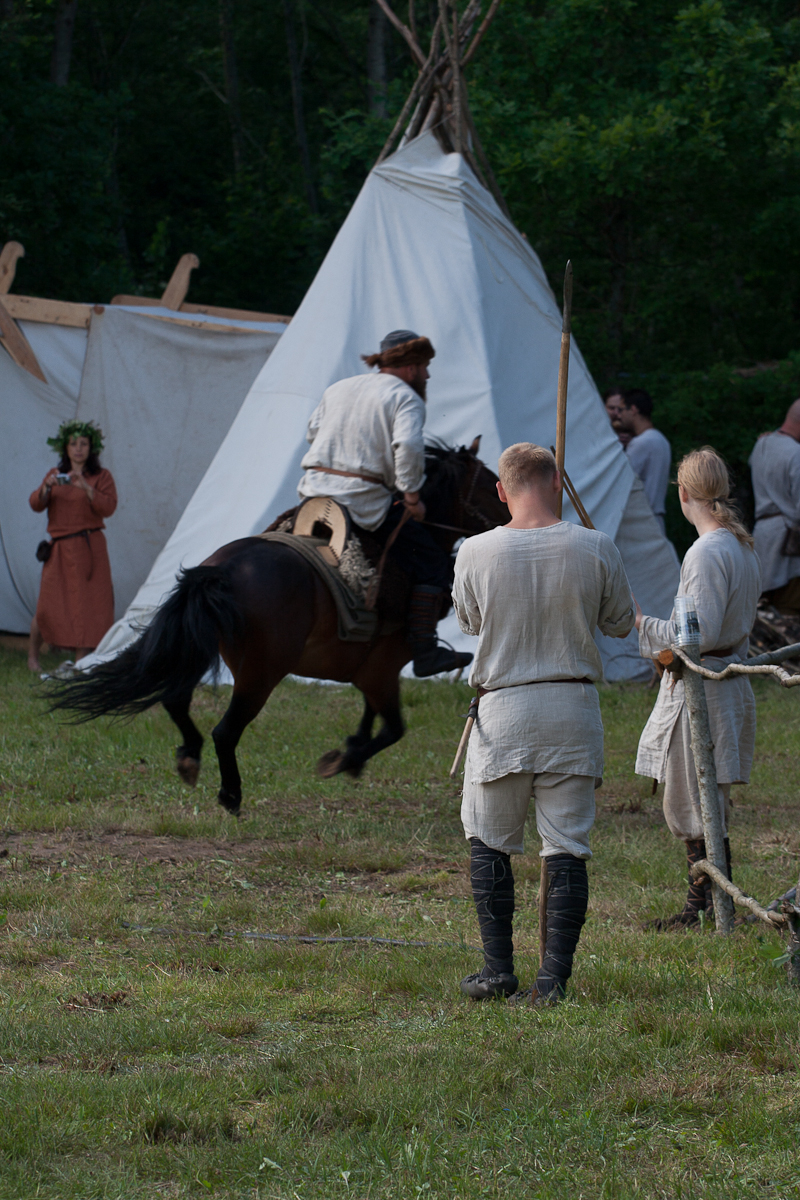 A scene of medieval reenactment at Kilkim Žaibu 2012 shows people dressed in period costumes. In the foreground, two men in beige tunics and boots stand, one holding a spear. A man on horseback rides past them. In the background, a woman in a long dress stands near a white canvas tent. Green trees surround the area.