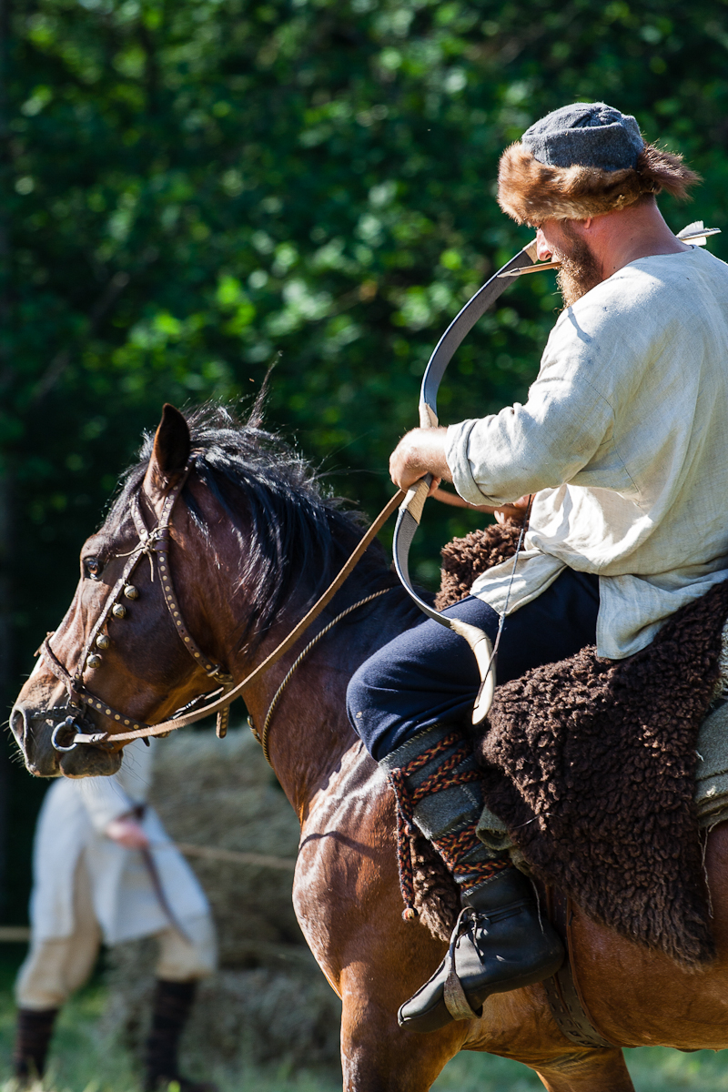 A man dressed in medieval attire, including a fur hat and a light tunic, is riding a brown horse. He holds a curved bow, ready to shoot. The scene, reminiscent of Kilkim Žaibu 2012, unfolds in a lush, green forest with another person in similar attire visible in the distance, partially obscured.