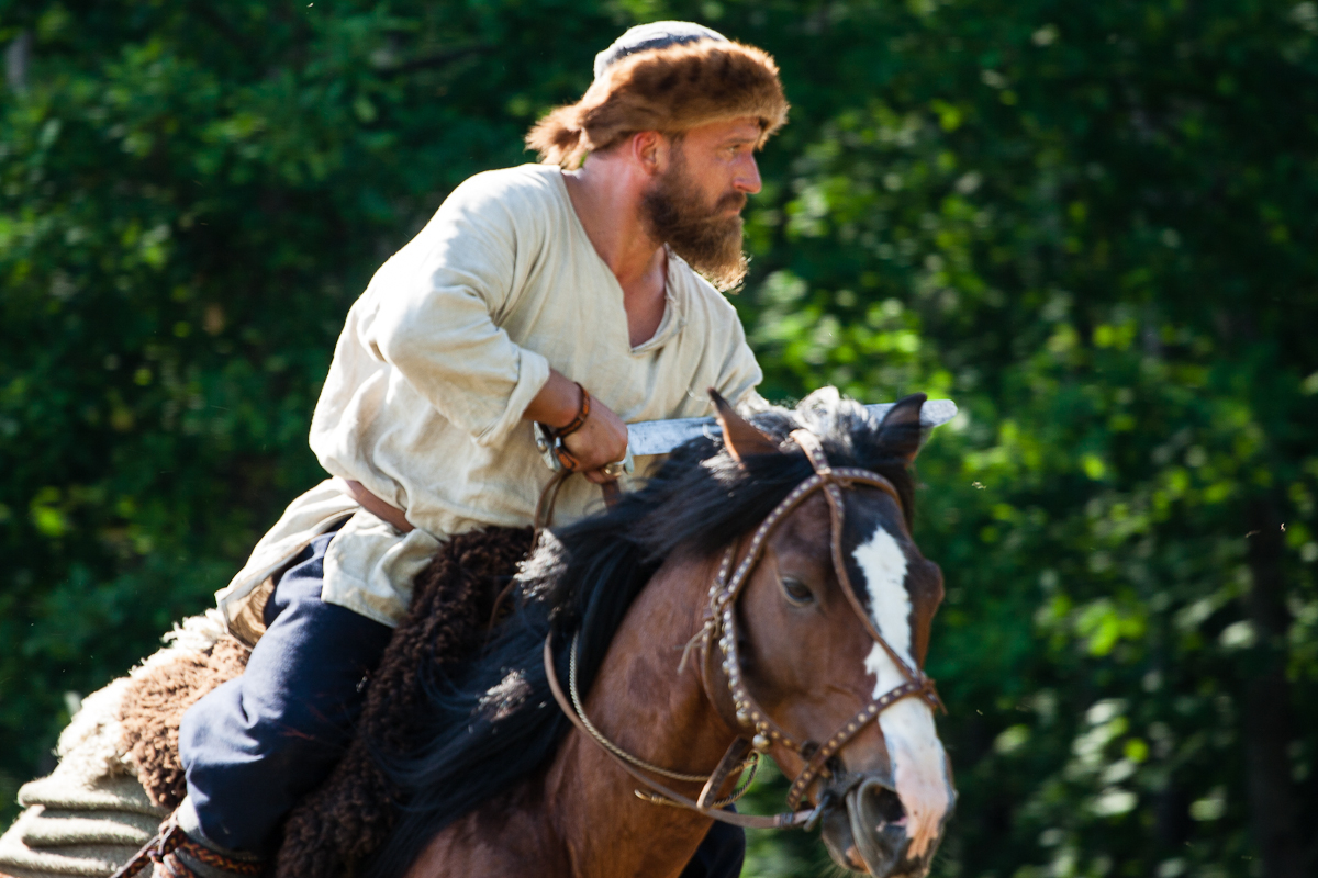 A man dressed in medieval attire with a fur hat and white tunic rides a brown horse with a white stripe on its face. He looks focused and determined, holding the reins with both hands. The background, reminiscent of Kilkim Žaibu 2012, is a lush forest with dense green foliage, perfect for the festival vibe.