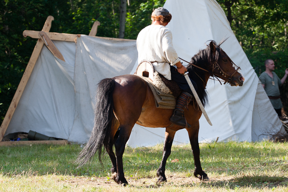 A person dressed in historical attire rides a brown horse in front of a white canvas tent with wooden supports in a grassy, wooded area at the 2012 Kilkim Žaibu festival. The rider's back is turned to the camera, and other people and trees can be seen in the background.