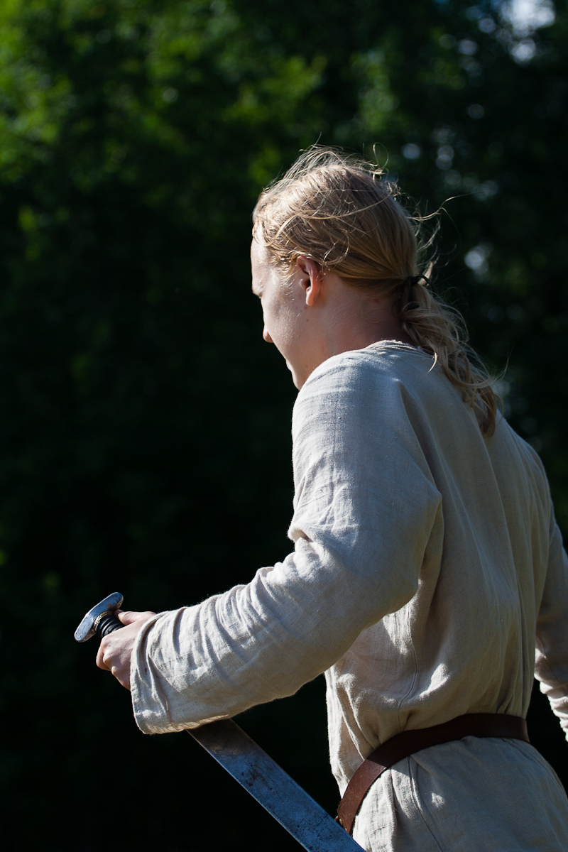 A person with long, light hair tied back, wearing a light-colored tunic, stands outdoors with trees in the background at Kilkim Žaibu 2012. The individual holds a sword in their right hand, the blade pointed downwards. The scene is set in daylight, highlighting the person's back and right side.