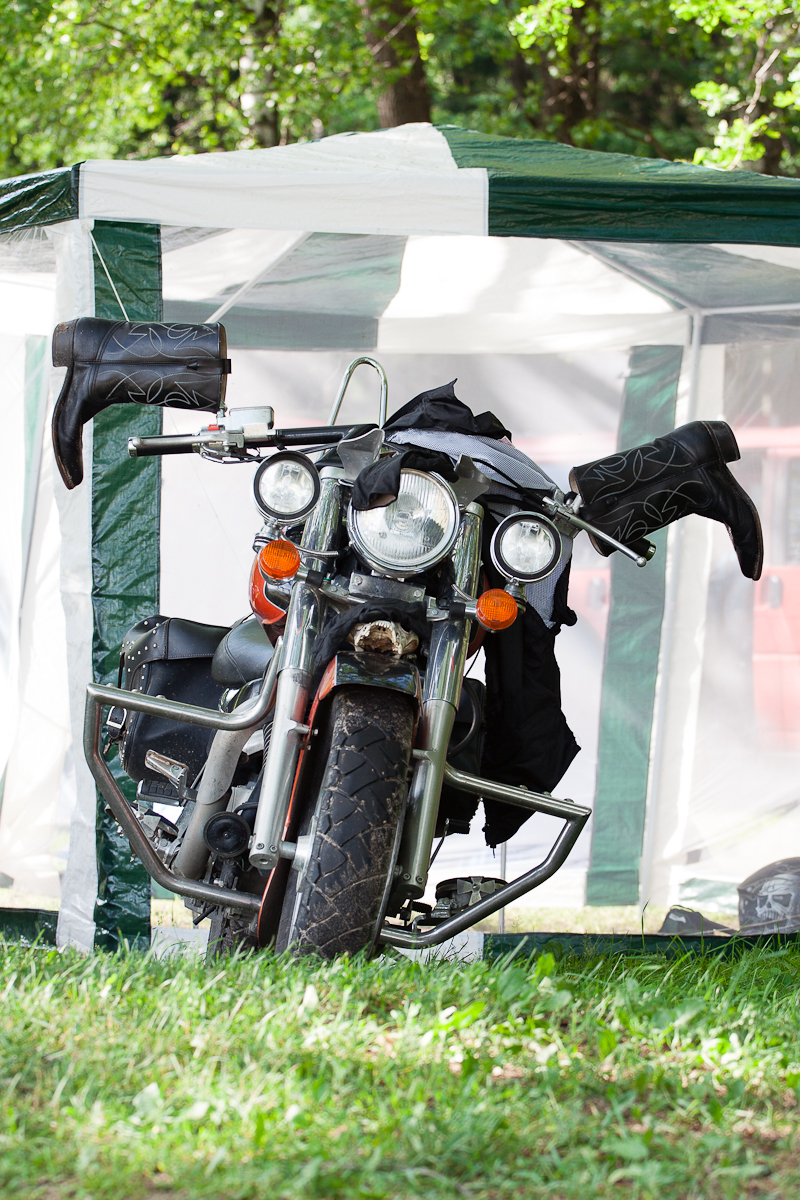 A motorcycle is parked on grassy ground inside a white and green canopy tent at the Kilkim Žaibu festival. The bike is partially covered with a black jacket draped over the handlebars, and black motorcycle chaps hanging from either side of the tent frame. The background shows green foliage and a red vehicle.