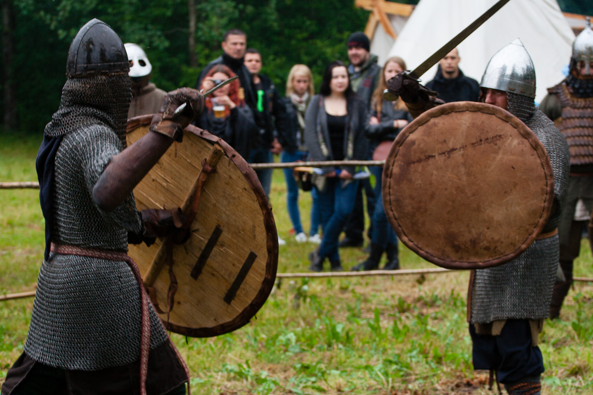 Two people clad in medieval armor and carrying large round shields engage in a mock sword fight at the Kilkim Žaibu festival. The audience, dressed in modern casual attire, watches intently. The scene takes place outdoors with greenery and a rustic tent visible in the background, evoking the spirit of 2012.