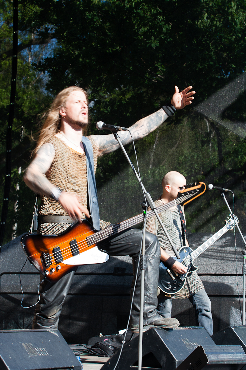 Two men perform on a stage at Kilkim Žaibu 2012, both wearing chainmail armor. The lead performer, with long hair and tattoos, energetically sings into a microphone with his left arm raised. He also plays a bass guitar. The other man, bald, plays an electric guitar in the background. Trees are visible outdoors.