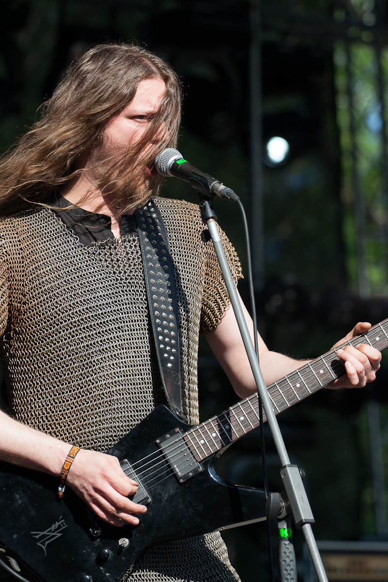 A musician with long hair performs on stage at the Kilkim Žaibu festival, wearing a metal chainmail tunic. He plays a black electric guitar and sings into a microphone. The background shows stage lighting and equipment, lit by natural daylight, suggesting an outdoor event in 2012.