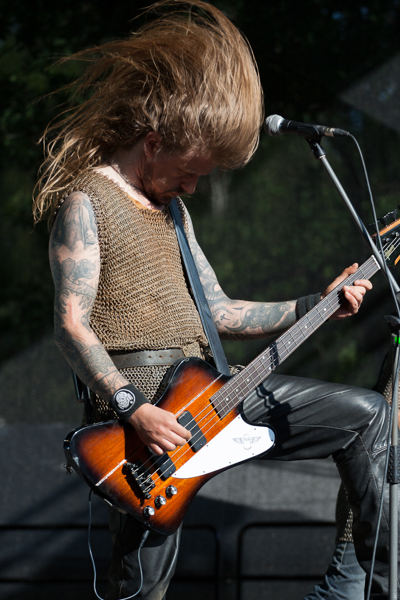 A long-haired musician, wearing a sleeveless chainmail shirt and black leather pants, energetically plays an electric bass guitar on stage at Kilkim Žaibu 2012. His head is down, hair flying mid-air in a dynamic scene. Tattoos cover his arms, and a microphone stand is positioned to his right.